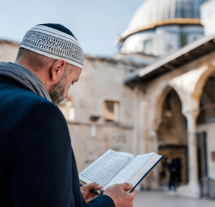 Muslim man reading the Qur’an with a mosque in the background at sunrise or sunset